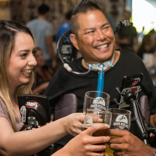 A group of people in a restaurant make a toast with beer glasses. Visible in the image are a blond, Caucasian woman and a tanned male wheelchair user who also has a vent tube at his neck. They are smiling and appear to be enjoying themselves.