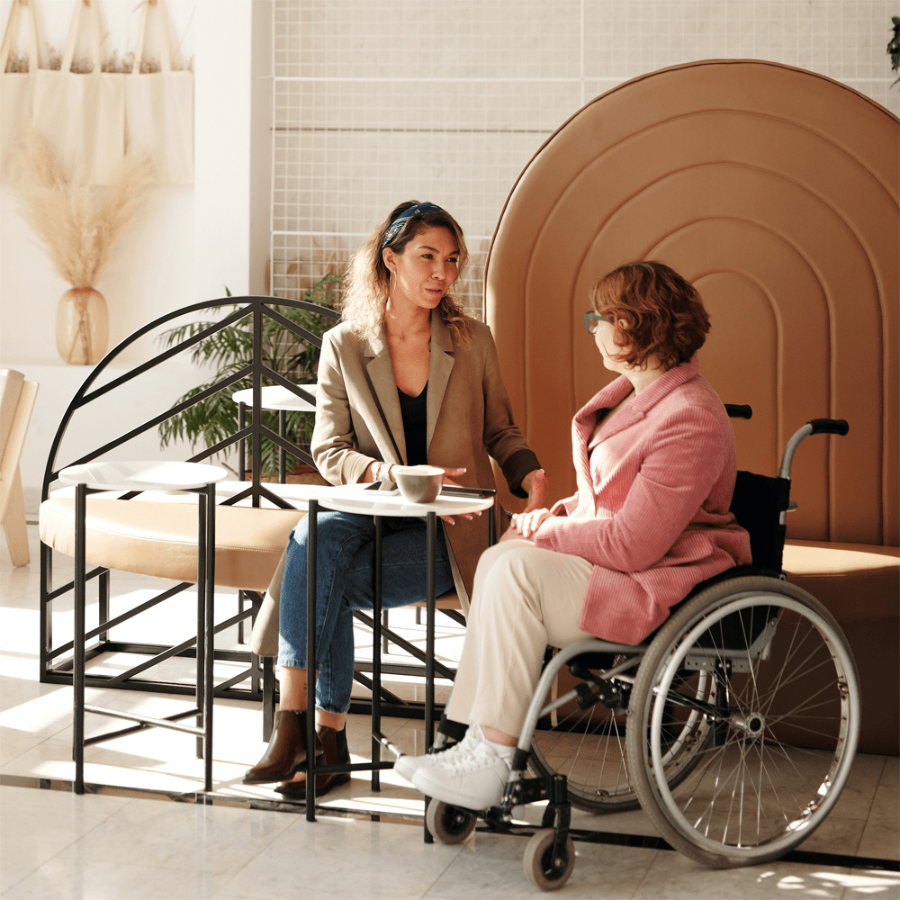 Two women are deep in conversation at a cafe. One woman sits on a bench seat and the other woman is seated in a wheelchair.