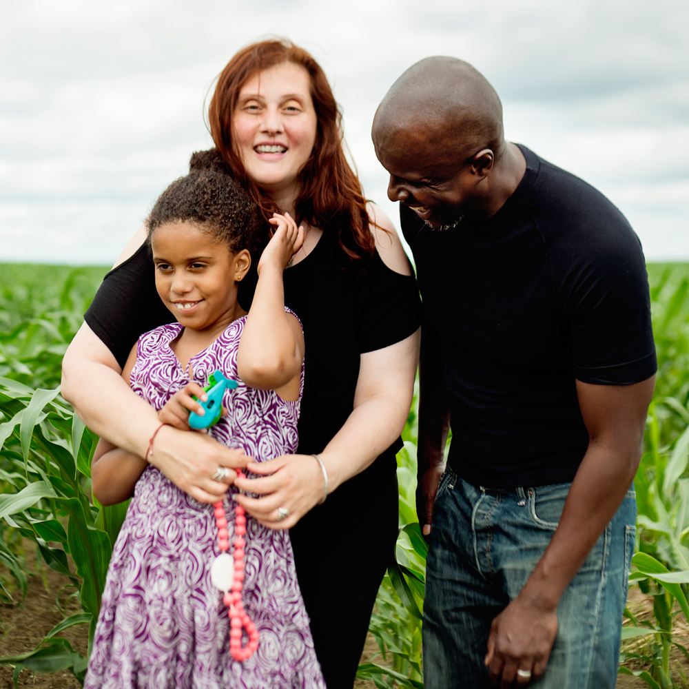 A family of three in an outdoor setting. The mother, who is Caucasian, has her arms casually wrapped around her school-aged daughter, who is holding a fidget. The father, a Black American man, is bending toward their daughter with a smile on his face.