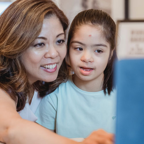A Latina mother and her daughter with Down syndrome look at a computer screen together. The mother is smiling and pointing to the screen and the daughter looks as if she is about to as a question.